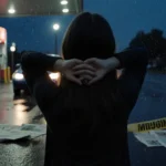 Missing teenage girl stands with hands behind neck at rainy gas station with dark sedan headlights glowing on wet pavement