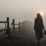 Katie Bates walking alone in morning fog with wilting flowers in hand and broken fence revealed in distance.
