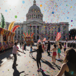 Floats parade with vibrant banners and marching band in sunlit streets, people dancing.