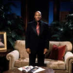 Montel Williams stands before worn beige couch with lush greenery and provocative magazine visible on table