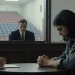 Mother sits with bowed head and clasped hands at courtroom table with judge watching and school auditorium visible through wi