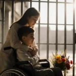Mother embracing son in wheelchair with prison fence and flowers on ground
