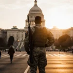 National Guard soldier stands watch with hand on rifle and Capitol dome glowing at sunset