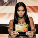 Nicki Minaj proudly holding a gold-plated credit card with the Trump Gold Card in front and a modern desk backdrop.