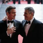 Noah Wyle and George Clooney standing together on red carpet with hands touching and flashbulbs glowing behind them