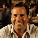 Norm Peterson smiling warmly at the bar with Cheers patrons talking behind him and golden lighting highlighting his friendly