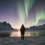 Person standing by frozen lake watching Northern Lights with snow mountains and golden sunset glow