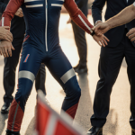 Hands holding up altered suit cheating attempt with Norwegian ski jumping officials reacting and a red flag nearby