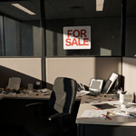 Office cubicle filled with clutter and a for sale sign in glass wall with natural light pouring in