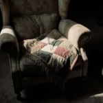 Old armchair holds faded patchwork blanket with cat searching floor nearby in dim nostalgic room