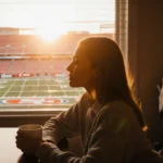 Olivia Culpo sits with coffee gazing out window with 49ers stadium visible through blinds and baby gear nearby