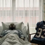 Osi Umenyiora lying still in hospital bed with NFL jersey draped nearby and medical equipment showing