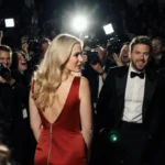 Pamela Anderson stands alone in red gown with Seth Rogen laughing behind her and paparazzi cameras flashing