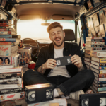 Pete Davidson holds up a VHS tape with a playful smile inside his Dad Van surrounded by tape stacks and warm light.