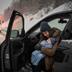 Mother cradling newborn in snowy Pittsburgh highway car with emergency responders and hospital headlights nearby