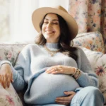 Pregnant woman relaxing on couch with pastel floral decor and floppy hat on wrist (91 chars)