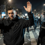 Protester raising hands in surrender with police presence and chaotic backdrop.
