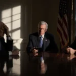 Three Republican senators sit tensely at polished wood table with one checking phone and shadow looming on wall