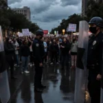 Riot shields stand with black tape covering pepper spray compartments near police uniforms and protesters with signs on rainy