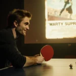 Robert Pattinson as Marty Supreme holds a ping-pong paddle with a knowing smile and faded movie poster behind him