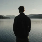 Rusty Yates stands by the lake with reflective water and melancholy expression showing grief