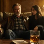 Sam Elliott sitting in armchair with whiskey glass and vintage photograph on table beside shadowed companion