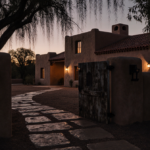 Stone path winds toward adobe mansion with terracotta roof and chimney on Santa Fe estate under dusk shadows.
