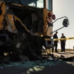 Crumpled school bus lies on rural roadside with shattered glass and emergency responders working near police car