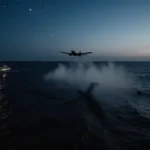 Search plane flies overhead casting shadow over waves with a lone boat lit at night and scattered debris on water
