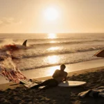 Injured surfer lies on Mendocino beach with cracked surfboard nearby and shark fin cutting through bloody water at sunset