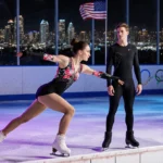 Female figure skater performing toe loop jump with floral outfit and Los Angeles skyline visible through frosty glass behind