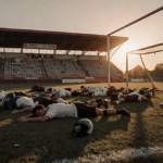 Injured bodies lying on a soccer field with scattered seats, shattered goalposts, a helmet under a setting sun