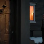 Federal agents stand near a worn wooden door with scratches and a peephole while a lit window casts orange light above