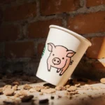 Starbucks cup with hand-drawn pig design sits abandoned on warm wooden counter with golden light highlighting its playful cha