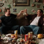 Adam Scott laughing with arms flailing while sitting on couch with Will Ferrell amid messy living room with snacks