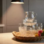 Colorful glassware sets stacked on woven tray with pendant light and fresh flowers creating café kitchen vignette