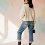 Woman modeling spring outfit with cropped jeans and cable knit sweater against pastel backdrop