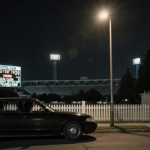 Sedan sits parked near picket fence under streetlight with stadium scoreboard and dark hair seen through rolled rear window.