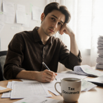 Person sitting at desk with coffee cup and organized Tax Documents folder in front while bills and calendar fill background