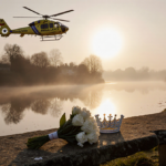 Helicopter hovering over riverbank with golden sunset reflection and memorial bouquet on bench.
