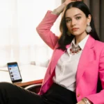Young woman wearing bright pink suit sits confidently at desk with smartphone showing TikTok comments and natural light strea