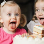 Toddler girl tasting birthday cake with wide eyes and pink dress while sister looks on with tongue out
