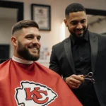 Travis Kelce getting a haircut with his barber Vince Garcia and wearing his Chiefs cape in warm barbershop lighting