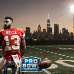 Travis Kelce stands with a football under his arm on a sunset field a spotlight highlights him over the Kansas City skyline.