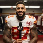Travis Kelce speaking into microphone with Chiefs jersey and locker room reflection behind