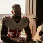 Travis Kelce sits with clasped hands near an ajar wooden door with a football field behind him and a vintage guitar leaning b