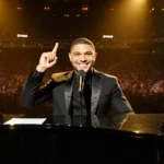 Trevor Noah playing piano with Grammy awards and microphone showing his confident smile