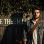 Tyler Hilton stands with guitar case and One Tree Hill sign visible through foliage
