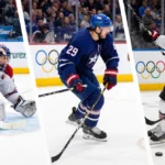 Auston Matthews scores a goal in ice hockey with Austen Ruesler defending and Olympic rings lit in the blur Italian backgroun