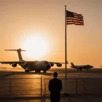 US military aircraft taxiing at dusk with American flag flying and shadowy figure watching near Qatar air base